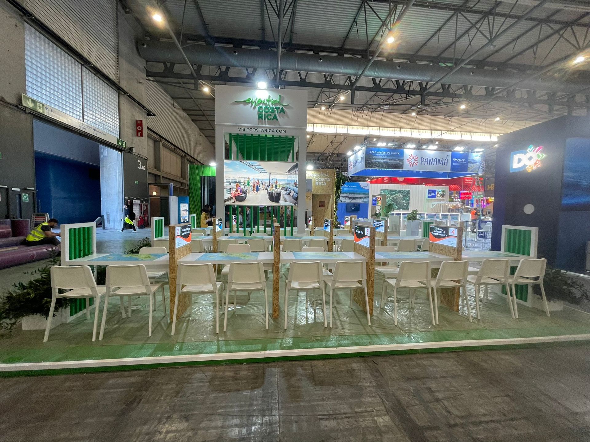 Un stand de exhibición con mesas y sillas, bajo una iluminación brillante. Detalles en verde y blanco.