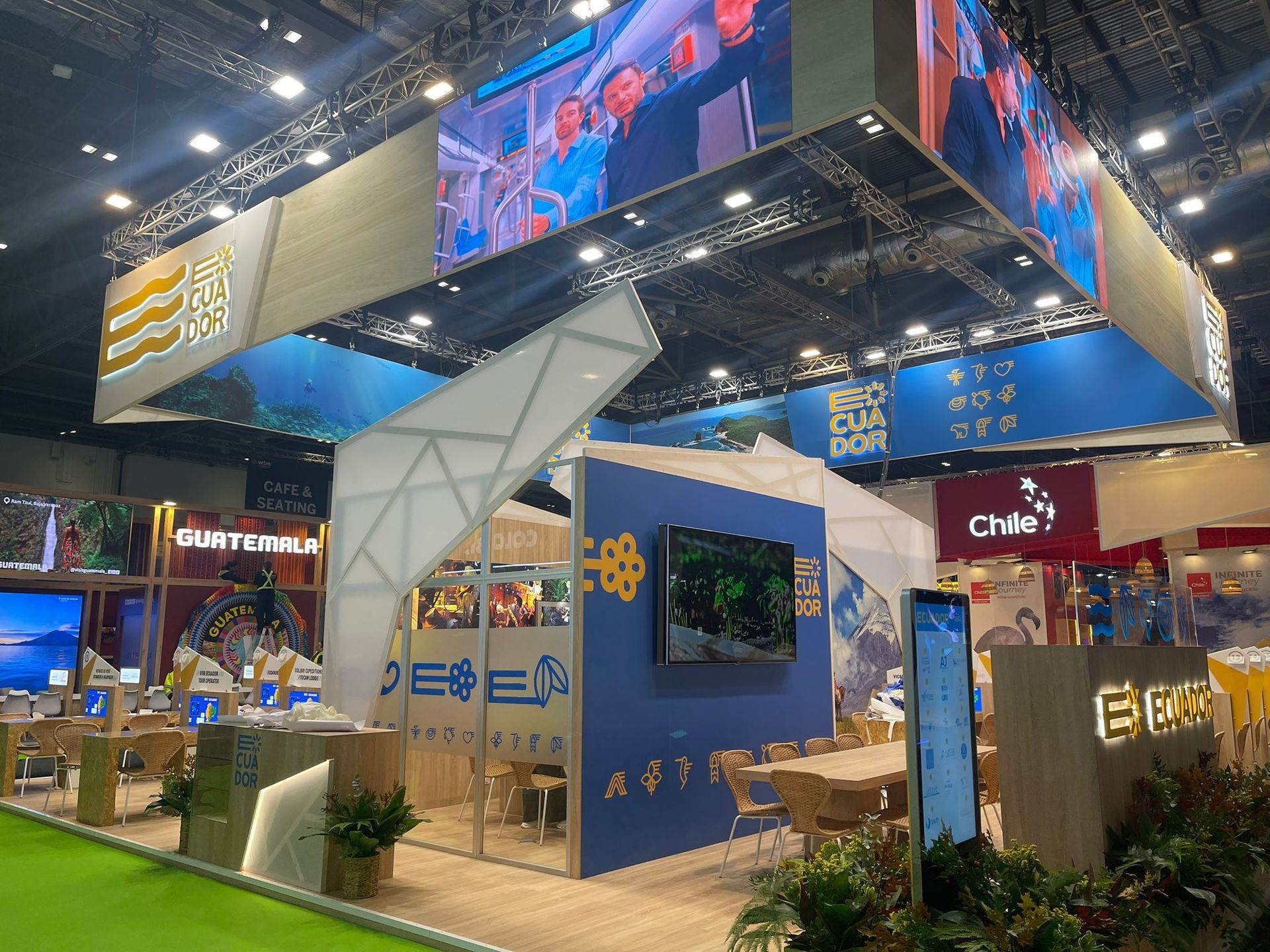 Un stand de exhibición con un diseño moderno, con pantallas, detalles en azul y amarillo y plantas en una feria comercial.