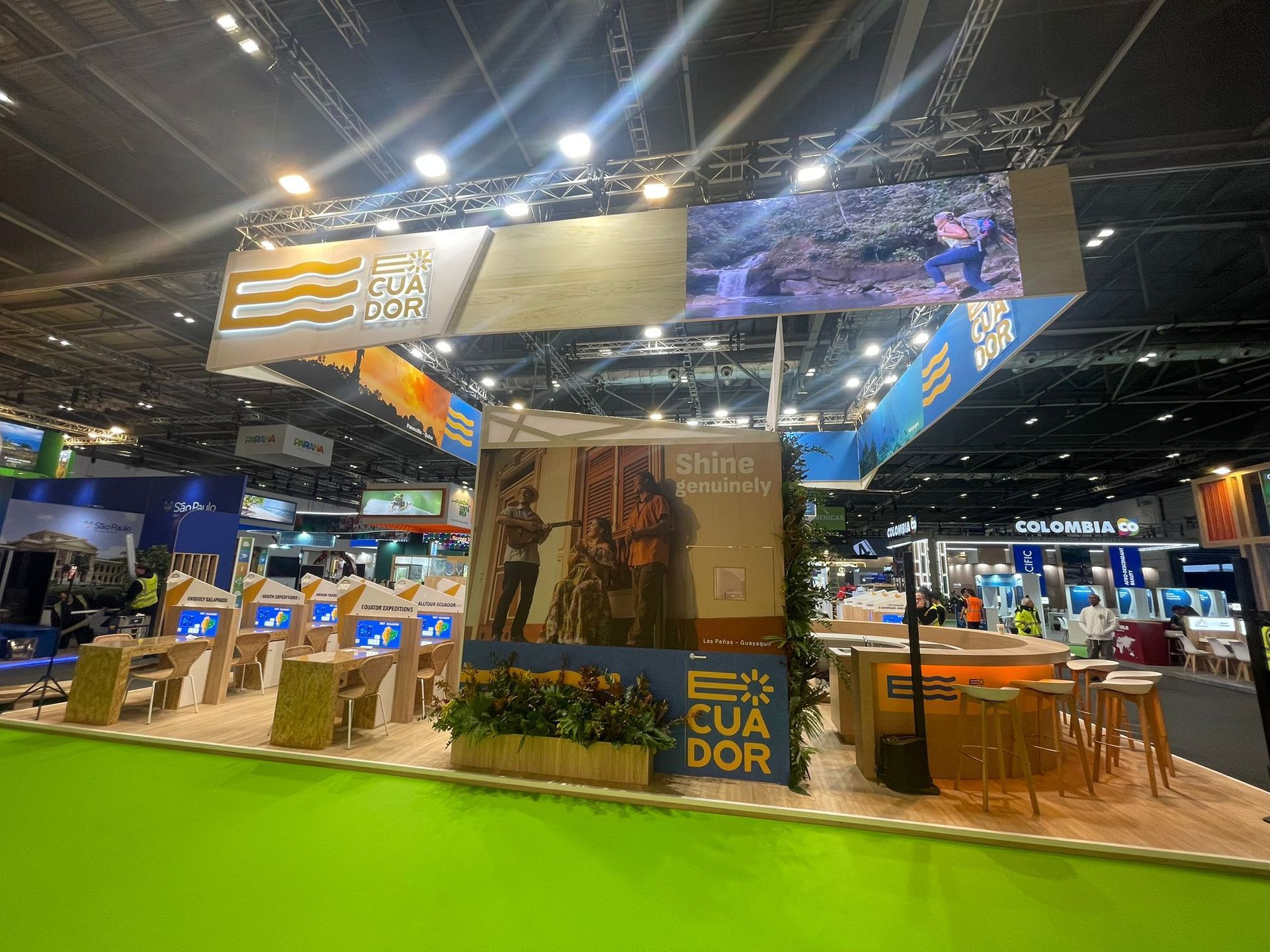 Stand de turismo de Ecuador en una feria comercial con expositores, señalización y gente. Alfombra verde, bien iluminada.