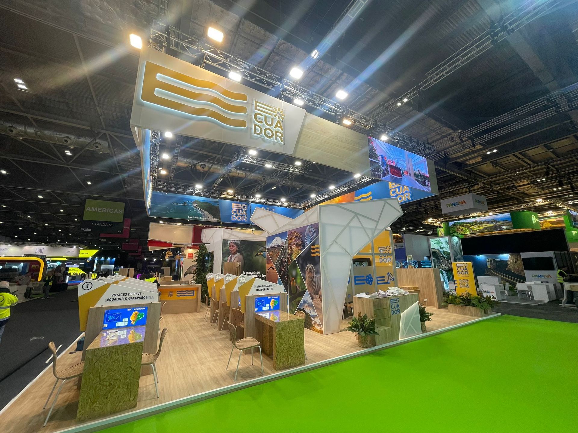 Stand de feria con detalles en amarillo y verde, que muestra información turística, con una alfombra verde brillante.