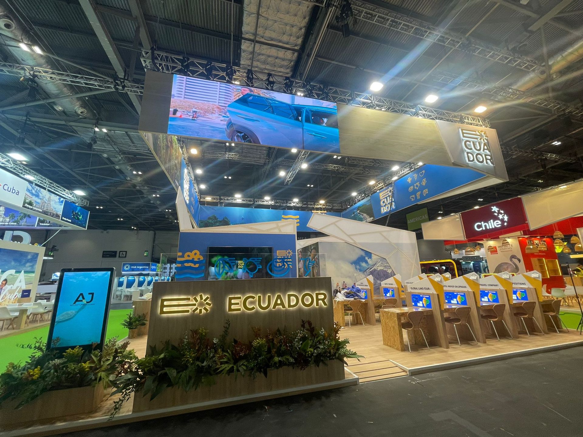 Stand de turismo de Ecuador en una feria comercial. Expositor azul y blanco con pantallas digitales, el logotipo de Ecuador y plantas.