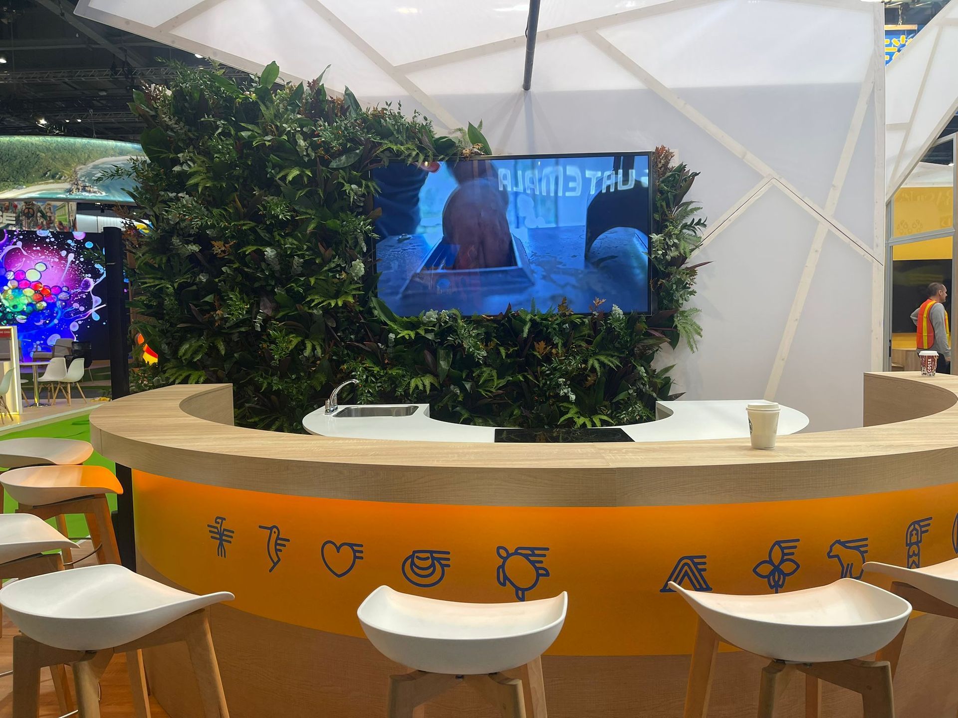 Área de bar con pantalla de TV, fondo de pared verde y taburetes blancos.