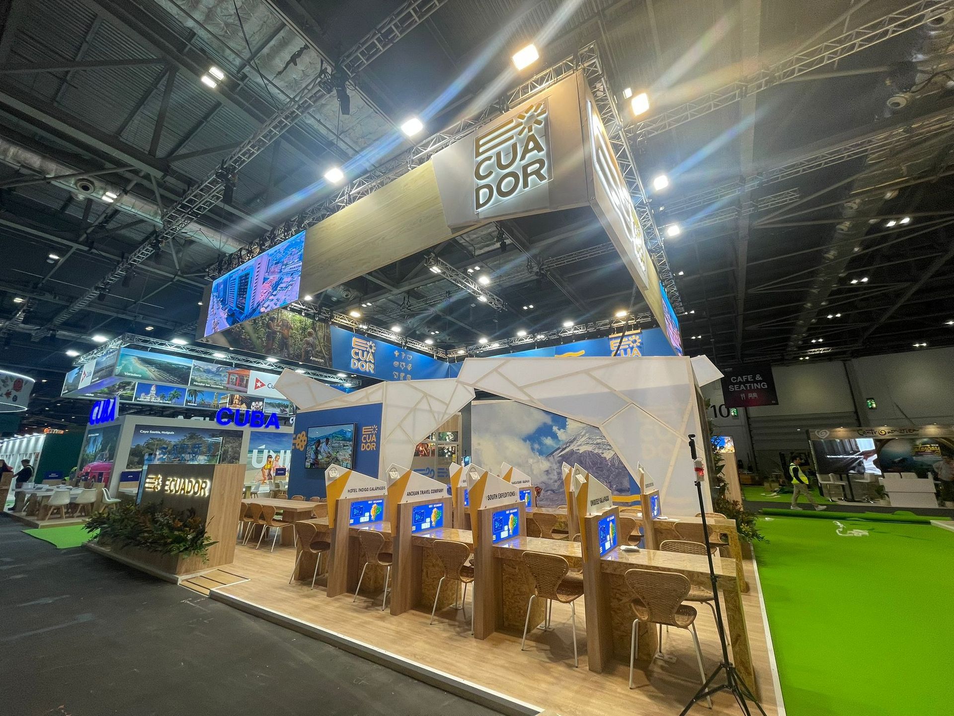 Stand de turismo de Ecuador en una feria comercial. Exhibidores blancos y azules, suelo color arena y monitores en los mostradores.