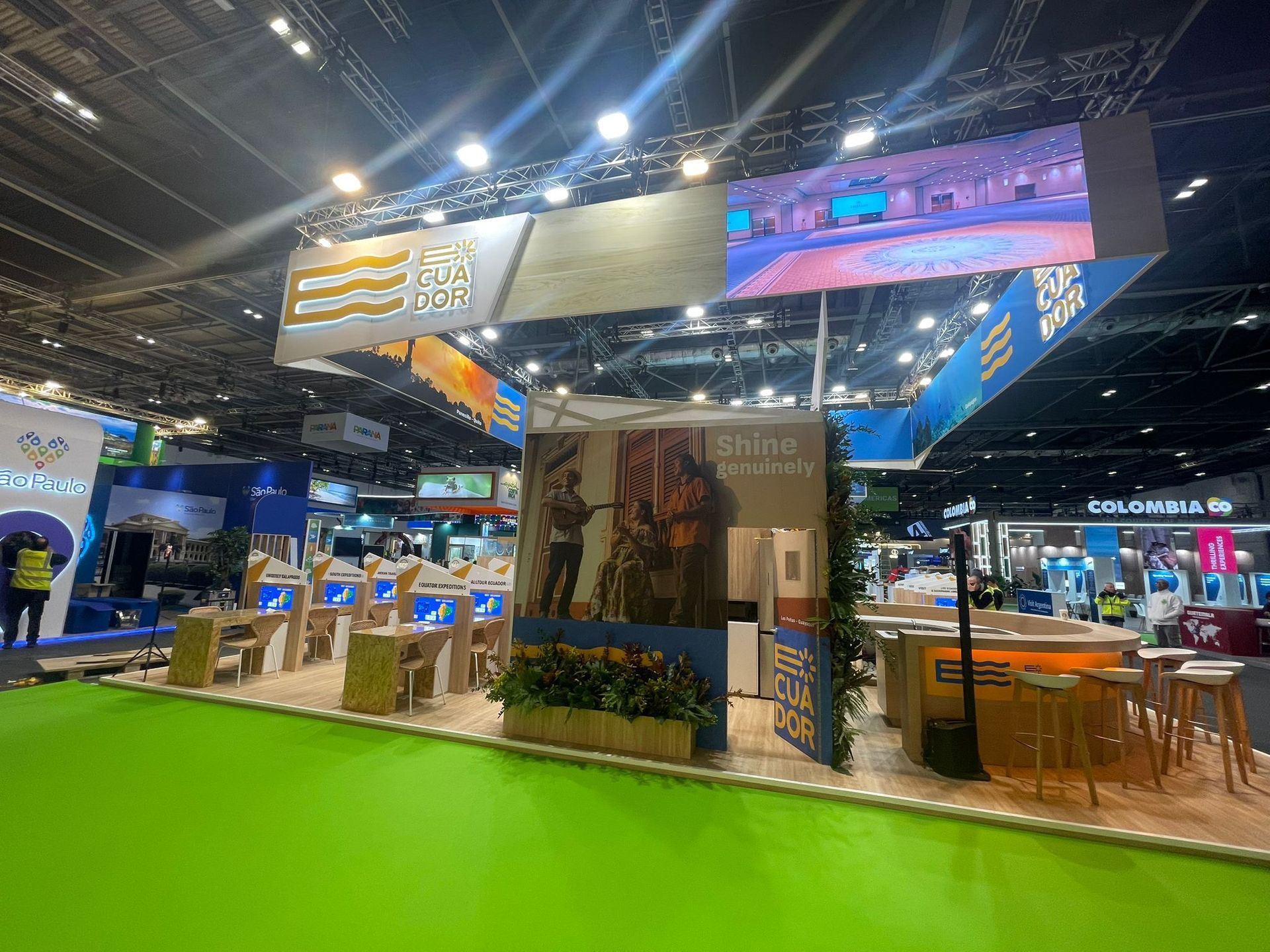 Stand de feria comercial con suelo verde, pantallas de visualización y zonas para sentarse.