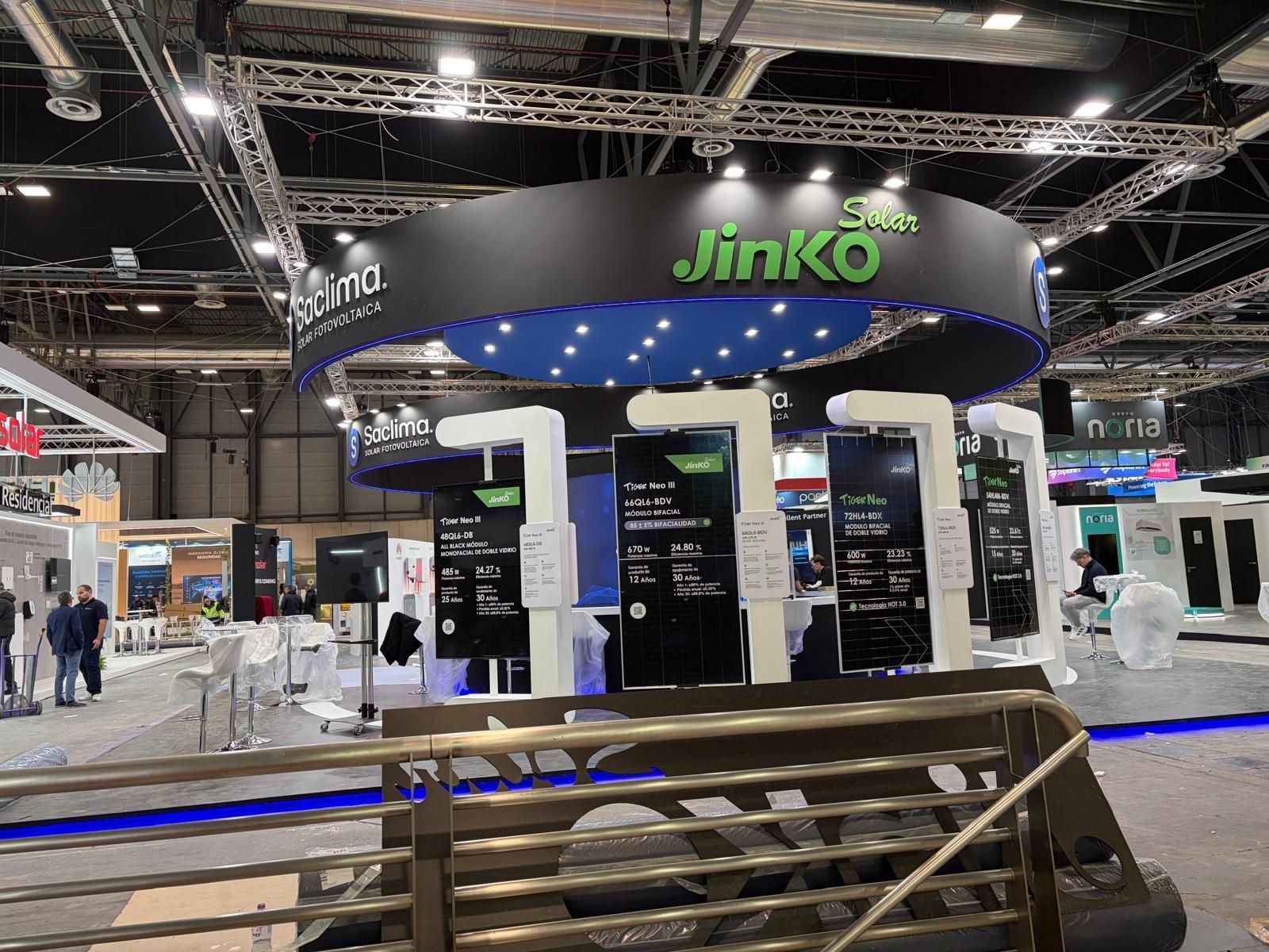 Stand de JinkoSolar en una feria comercial. Letrero circular negro con logotipo verde. Expositores con información. Iluminación brillante.