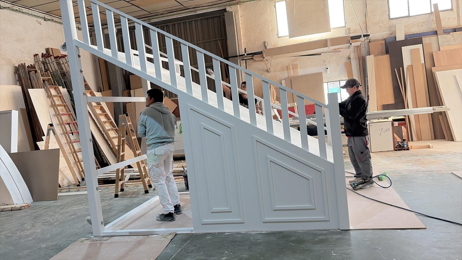 Dos personas instalando una escalera de madera blanca en un taller.
