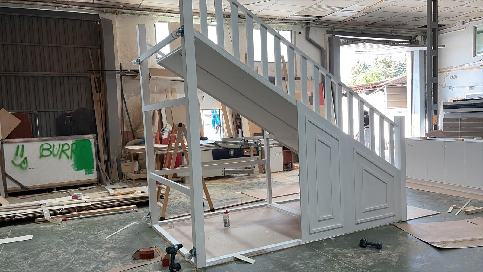 Escalera de madera blanca en construcción en un taller; barandilla, puertas de almacenamiento y marco expuesto.