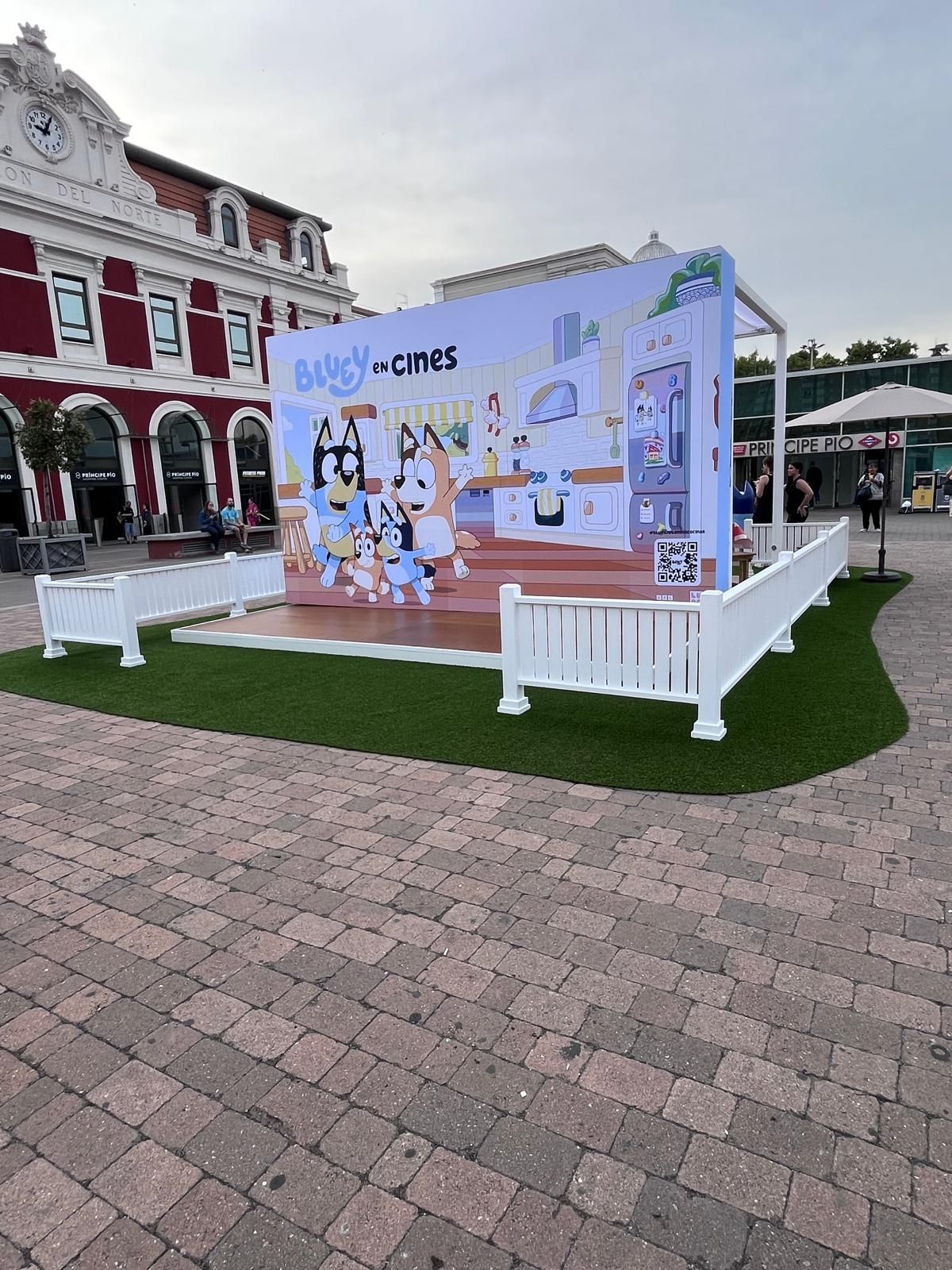 Escenario al aire libre con temática azul, con personajes de dibujos animados y una escena de cocina. Rodeado de una valla blanca.
