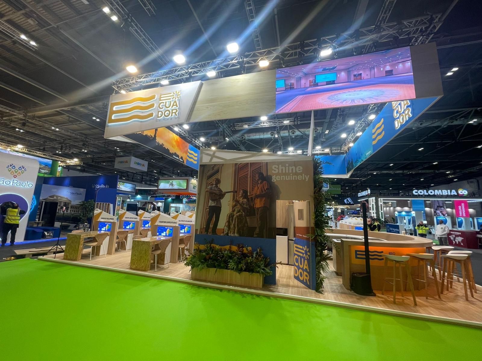 Stand de feria comercial con señalización, asientos y exhibidores; piso verde, varias marcas e iluminación brillante.