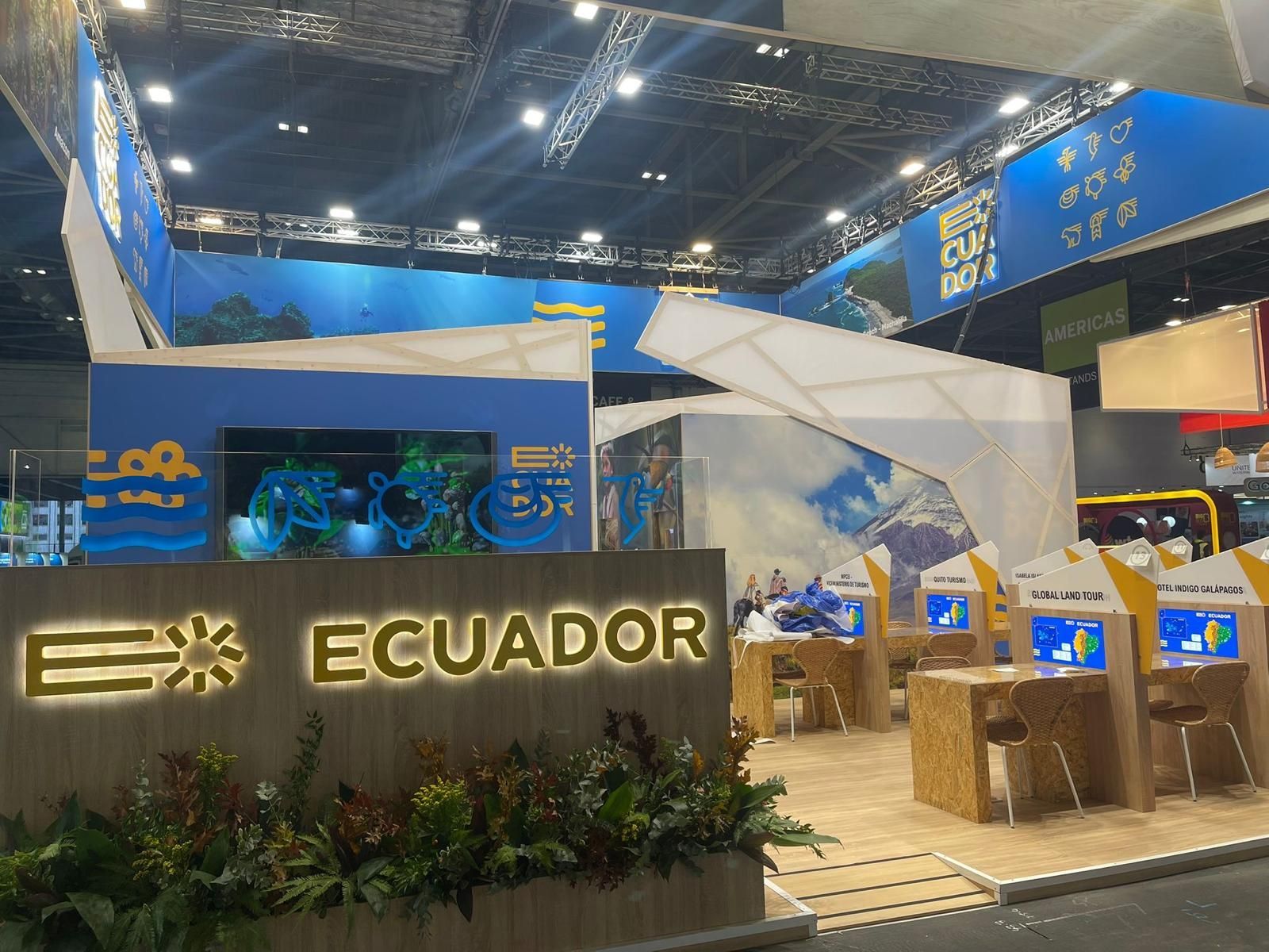 Stand de turismo de Ecuador con logo, decoración azul y blanca, plantas y displays interactivos en una sala de exposiciones.