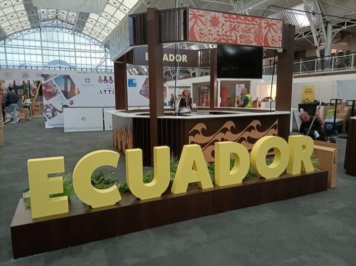 Stand de Ecuador en un evento con grandes letras amarillas, un mostrador y un televisor.