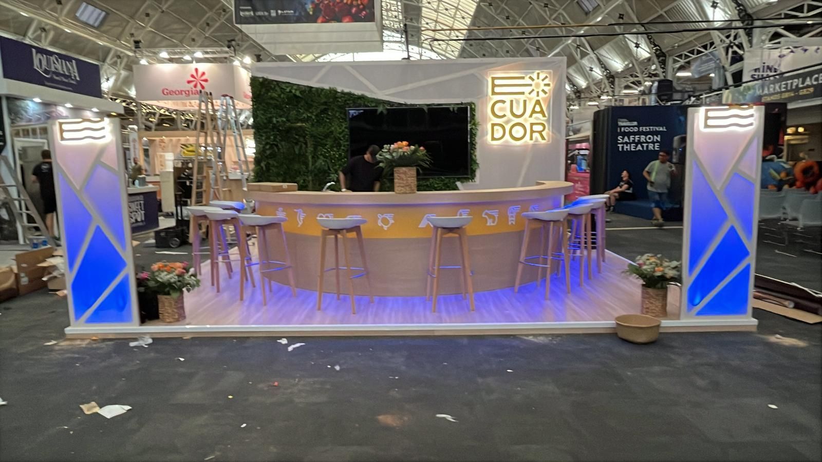 Exhibición de bar ecuatoriano en feria comercial: mostrador curvo, letrero iluminado, taburetes, pared verde y arreglos florales decorativos.