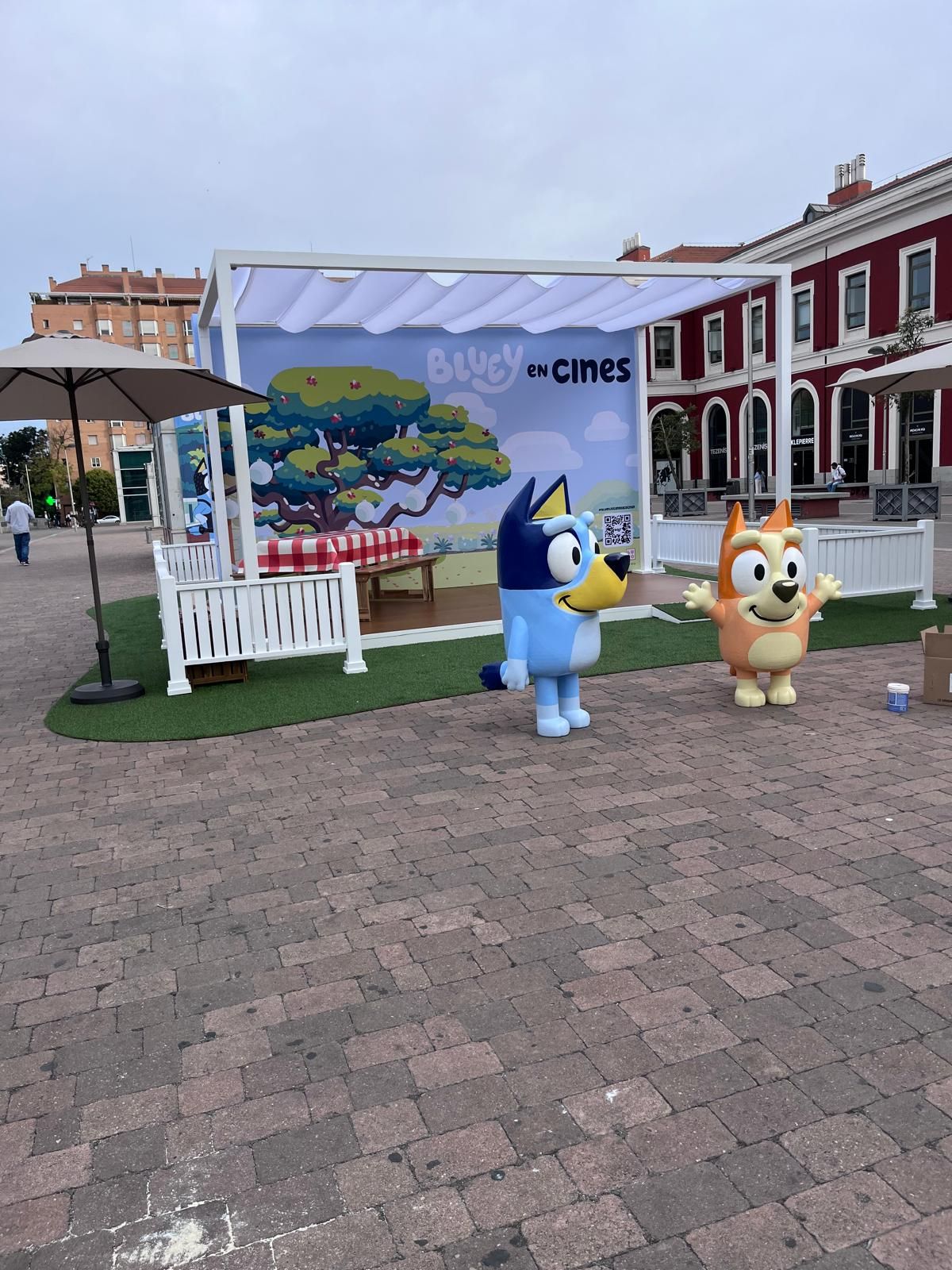 Los personajes de Bluey y Bingo aparecen en un evento promocional; escenario con pancarta y plaza de ladrillos rojos.