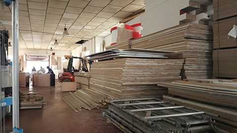 Interior de almacén con pilas de cajas de cartón y paneles de madera, carretilla elevadora, trabajadores.
