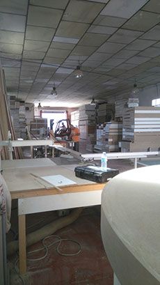 Interior de un taller; pilas de leña y mesas de trabajo. La luz entra a raudales por la puerta.