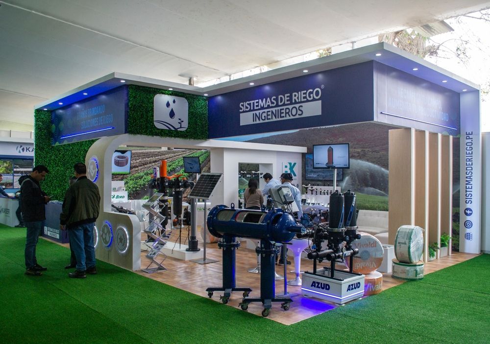 Stand de feria con sistemas de riego, con gente mirando equipos y señalización. Suelo de césped verde y detalles en azul y blanco.