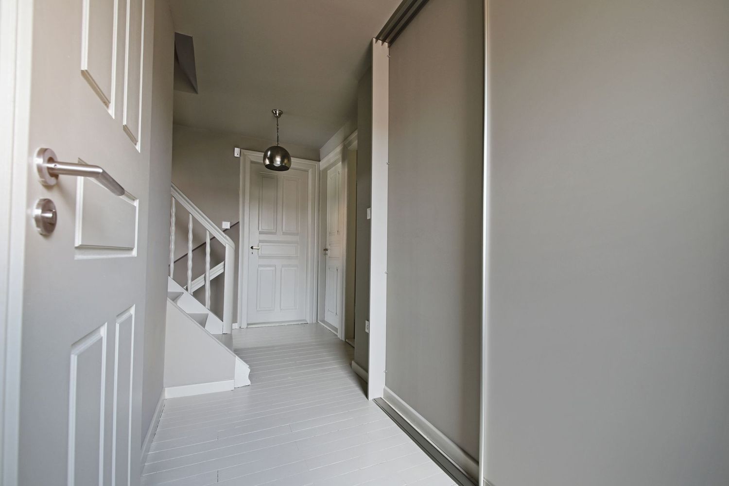 Couloir gris avec portes, escaliers et placard. Boiseries et sol blancs ; un seul luminaire est suspendu au plafond.
