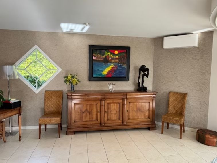 Salon avec armoire en bois, œuvres d'art, chaises et fenêtre en losange. Murs beiges, sol blanc.