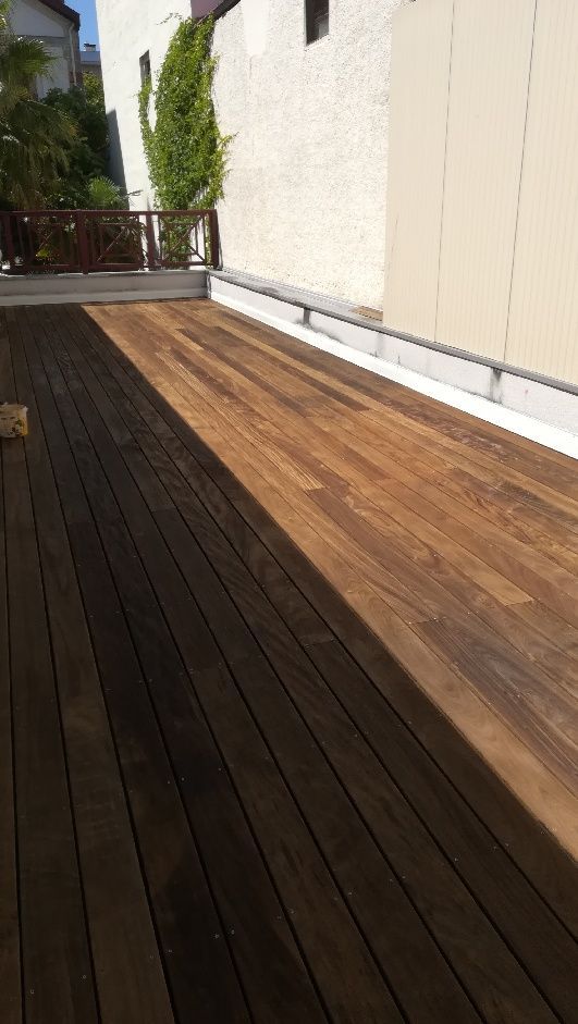 Terrasse en bois longeant un mur couleur crème, avec des planches foncées et une partie brune ensoleillée menant à une rambarde et à des plantes.