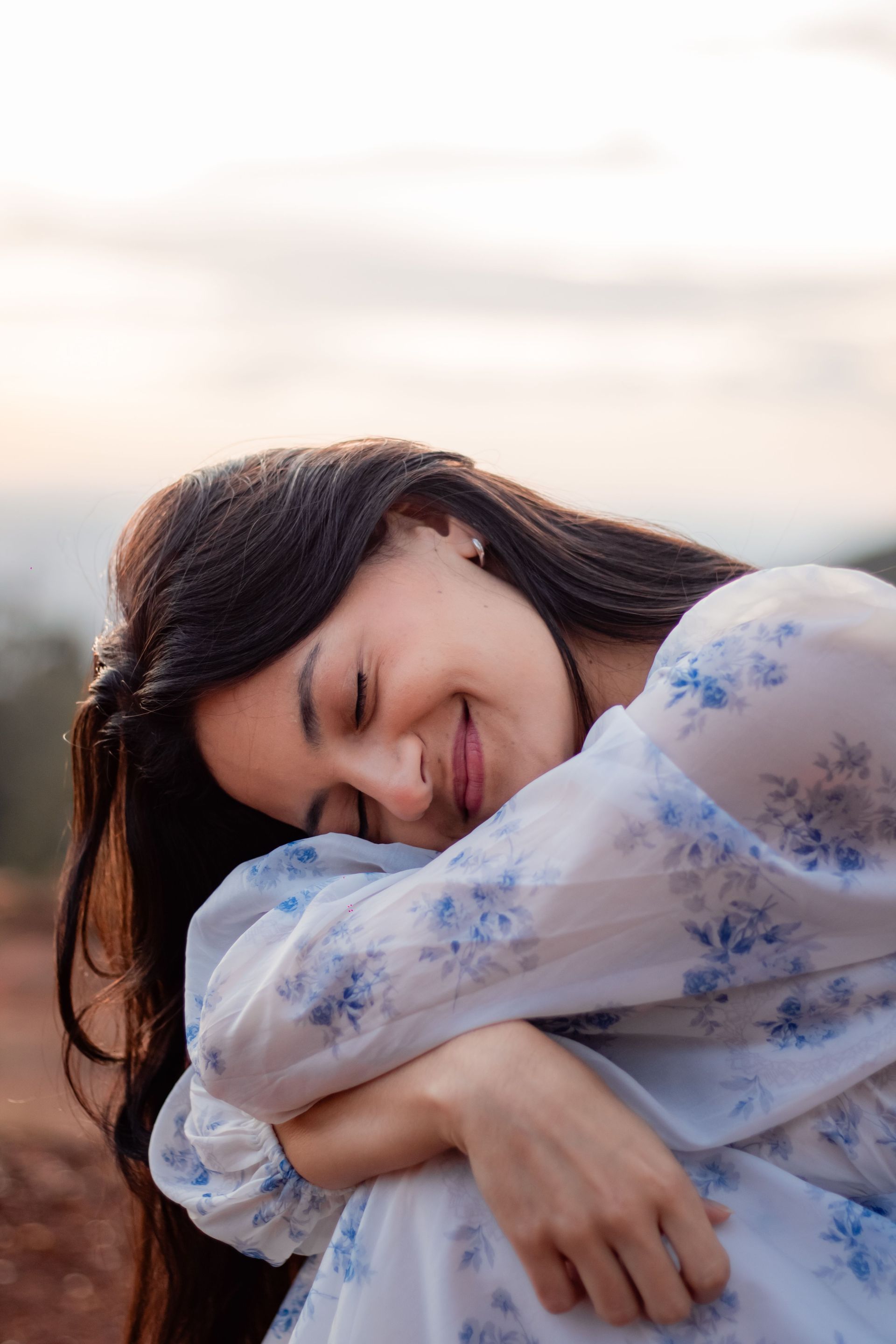 Mujer con la cabeza apoyada sobre los brazos cruzados, vestida con una blusa floral clara, con un suave cielo al aire libre de fondo.