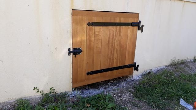 Trappe en bois avec charnières en métal noir, encastrée dans un mur blanc au bord de l'herbe.