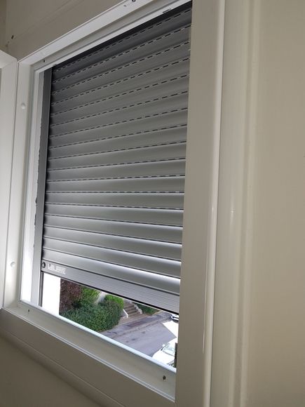 Volet blanc partiellement ouvert donnant sur une maison et de la verdure à l'extérieur