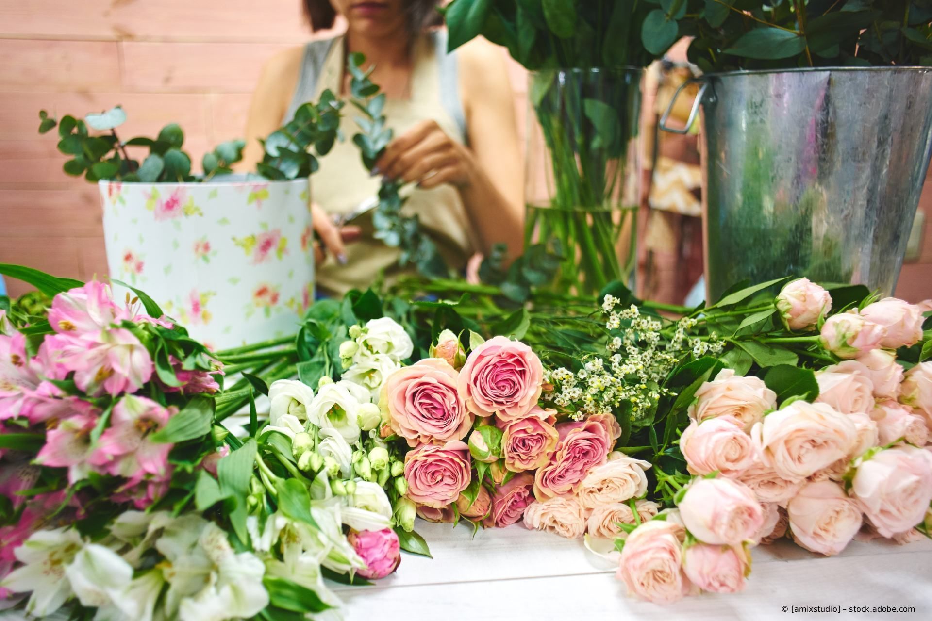 Eine Frau arrangiert Blumen auf einem Tisch mit verschiedenen Blüten und Grünzeug; die Szenerie ist eine Werkstatt.