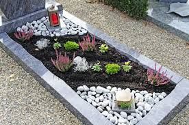 Grab mit dunkler Erde, verschiedenen Pflanzen, weißen Kieselsteinen und einer Kerze in einem grauen Steinrahmen.