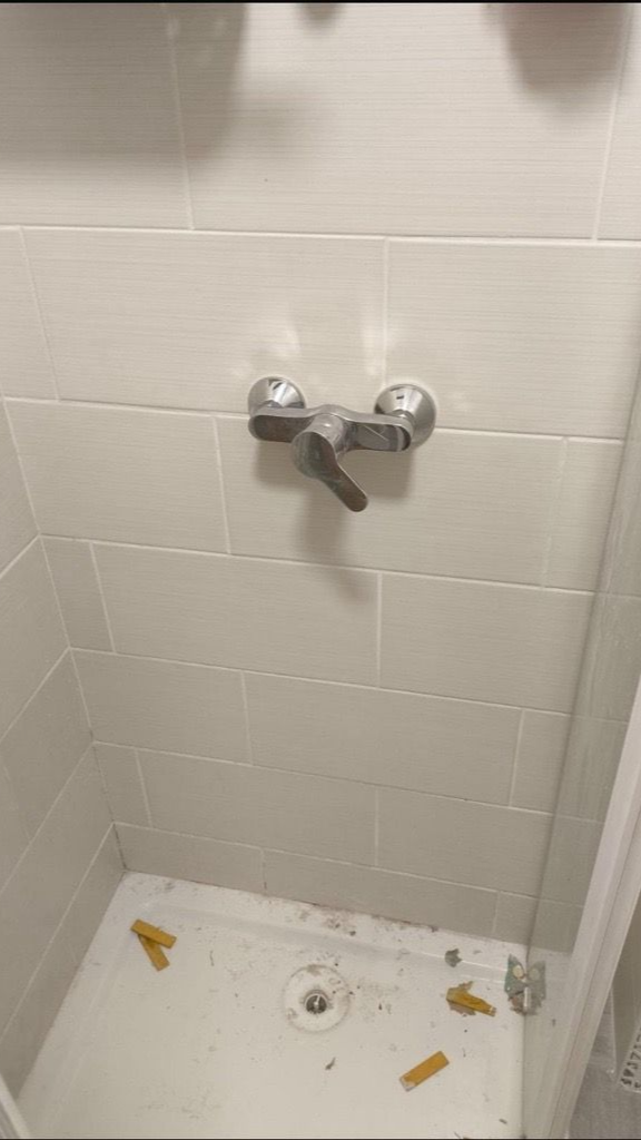 Cabine de douche avec robinet et bonde, murs carrelés gris clair, débris au sol.