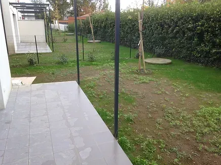 Terrasse avec sol carrelé et jardin gazonné avec poteaux métalliques et haie en arrière-plan.