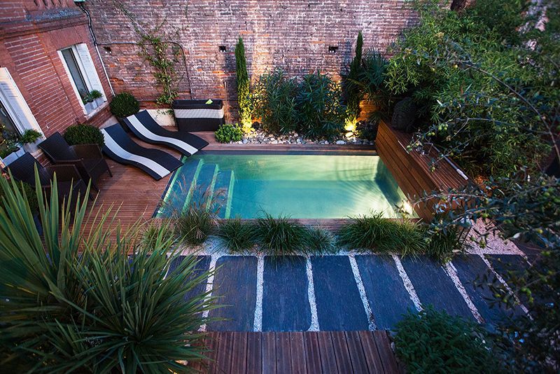 Photo d'une piscine au milieu d'une terrasse en bois avec de la verdure et des transats. 