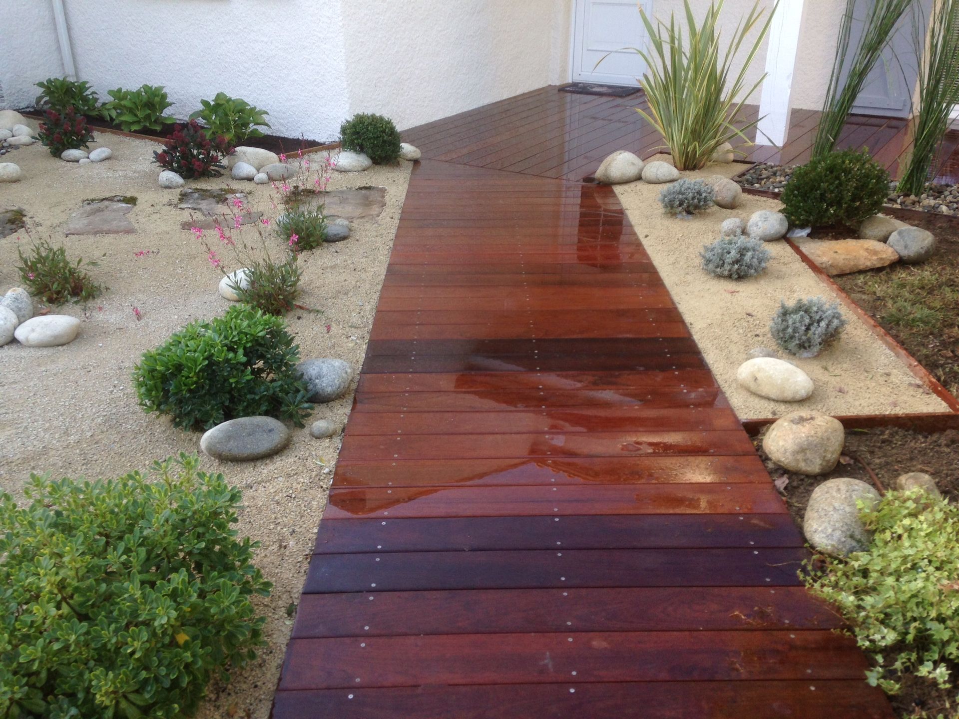 Photo d'une entrée de maison aménagée avec une allée en bois, des galets et un peu de verdure.