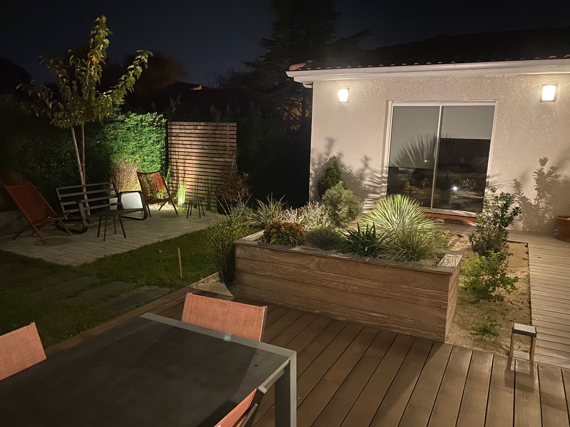 Vue nocturne d'un jardin avec une terrasse en bois, une table à manger et un parterre surélevé illuminé.