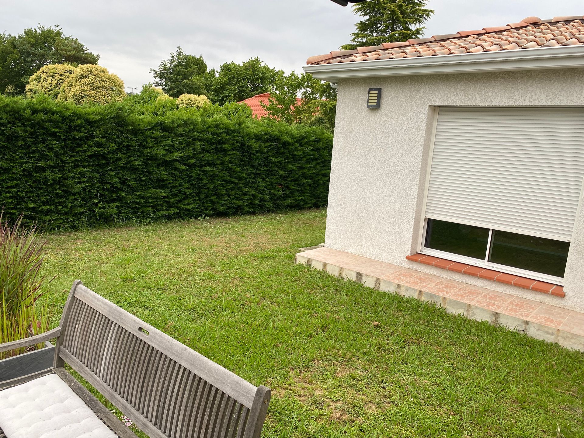 Pelouse, haie et bâtiment aux volets fermés. 