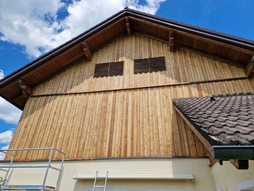 Holzfassade eines Hauses mit zwei offenen Fenstern unter einem braunen Dach vor blauem Himmel.
