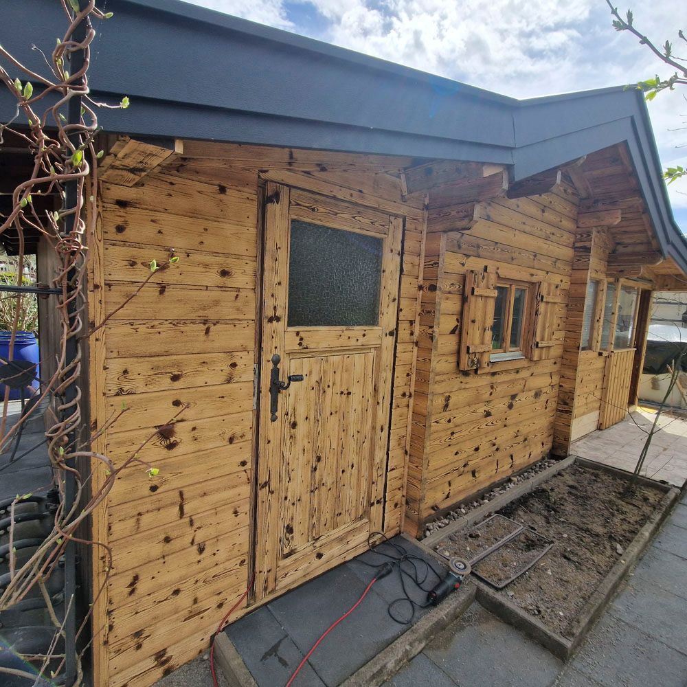 Ein Holzschuppen mit Tür, Fenster und kleinem Pflanzkasten unter blauem Himmel.