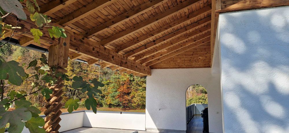 Ein Holzdach überspannt ein weiß getünchtes Gebäude mit einem Bogeneingang. Grüne Bäume und Sträucher sind zu sehen.