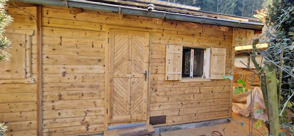 Holzhütte mit Tür, Fenster und Fensterläden, umgeben von Bäumen.