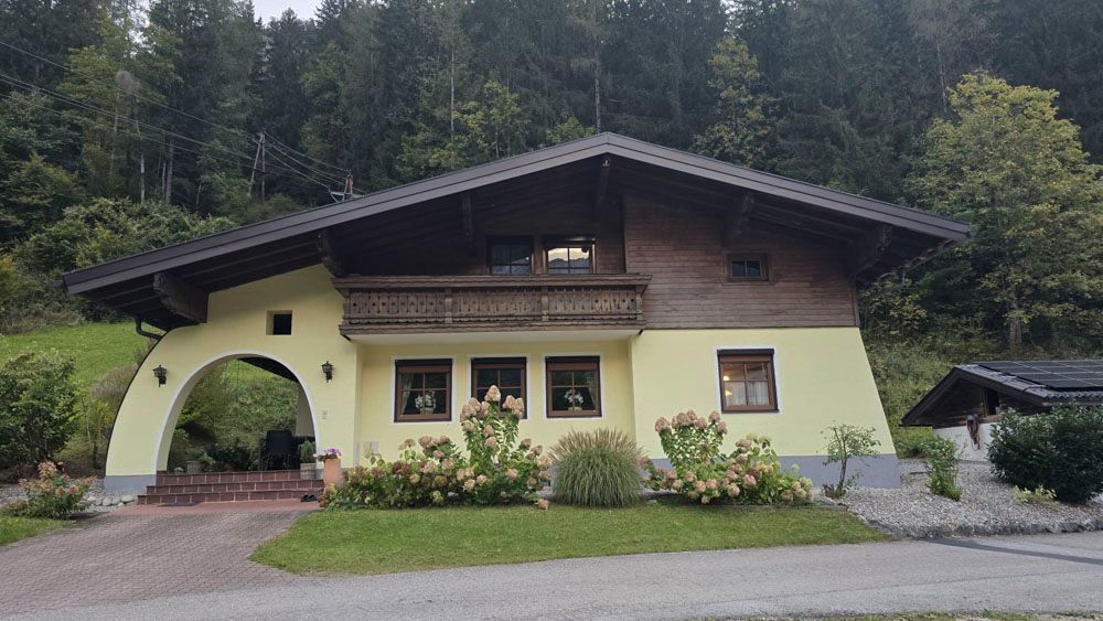 Gelbes Haus mit braunem Dach und bogenförmigem Eingang vor einem Wald.