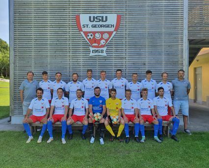 Die Fußballmannschaft der USU St. Georgen posiert in weißen Trikots mit roten und blauen Akzenten. Das Teamlogo ist sichtbar.