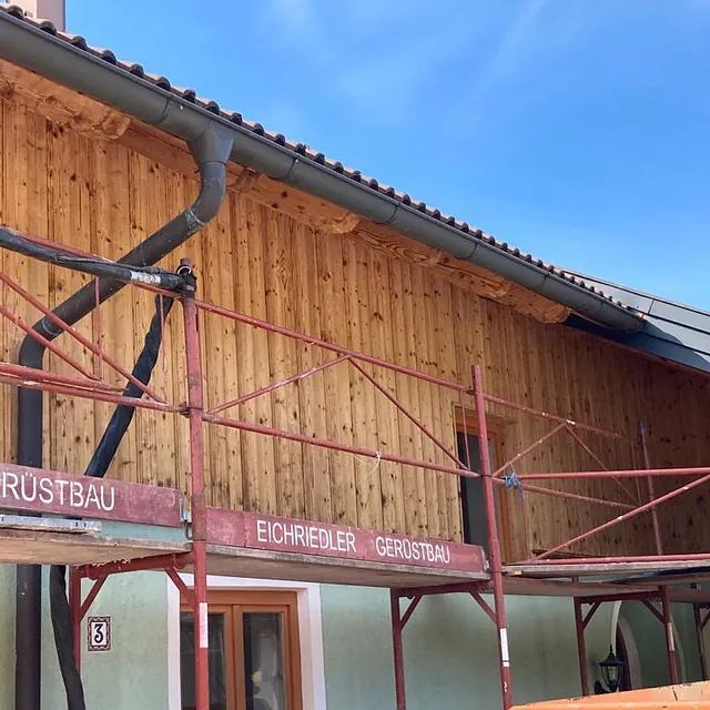 Ein Holzgebäude mit einem roten Gerüst davor, auf dem Rustbau steht