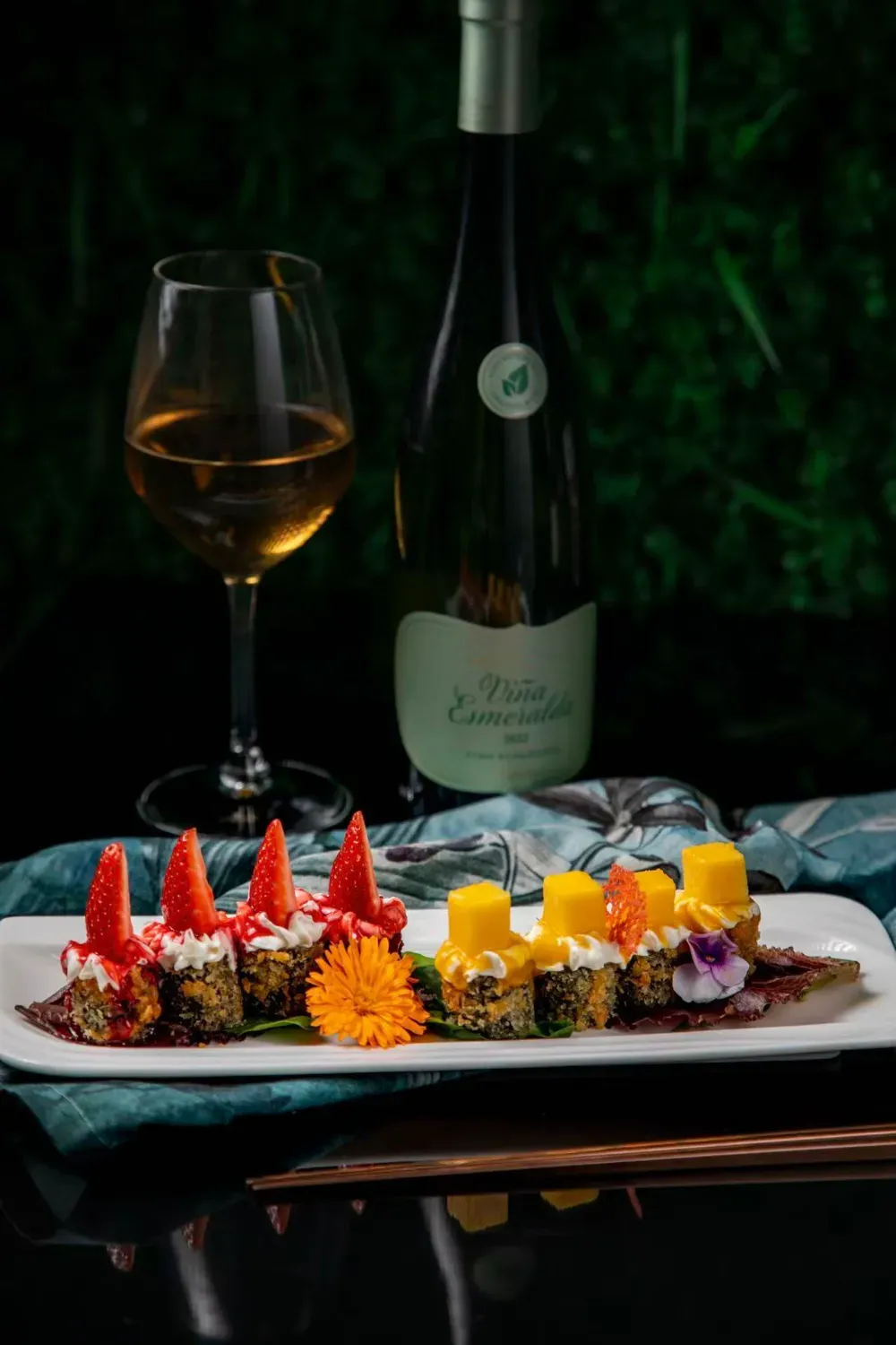 Un plato blanco alargado presenta sushi cubierto con rodajas de fresa y cubos de mango, junto a una copa