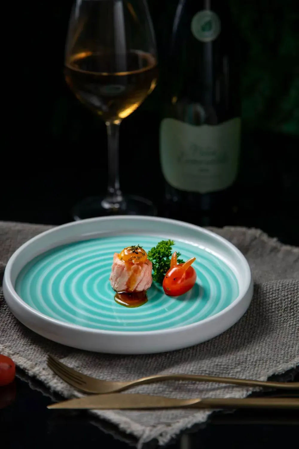 Un aperitivo de salmón con guarnición en un plato de color verde azulado con estampado en espiral, con una copa de vino
