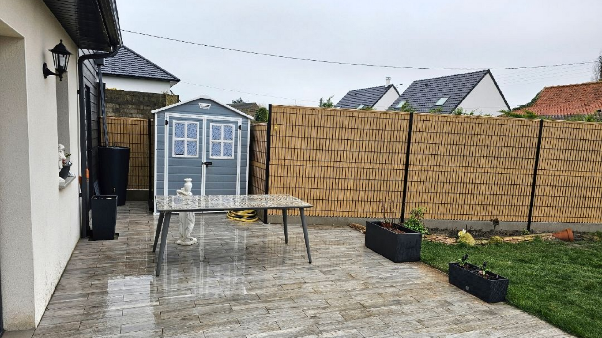 Terrasse avec une table, un abri de jardin et une clôture tressée marron sous un ciel couvert.