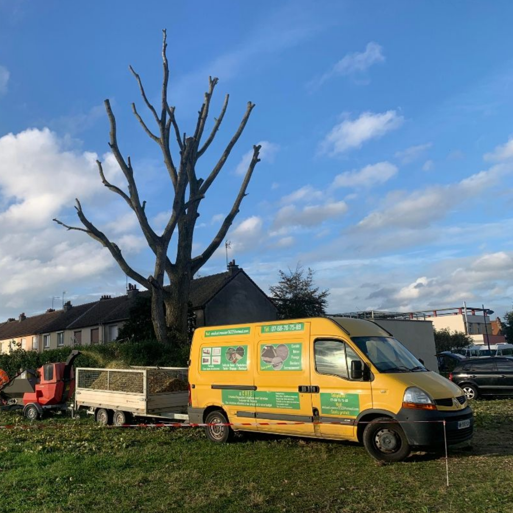 Une camionnette jaune avec remorque est garée à côté d'un arbre taillé dans un champ herbeux ; des bâtiments résidentiels se dressent à l'arrière-plan.