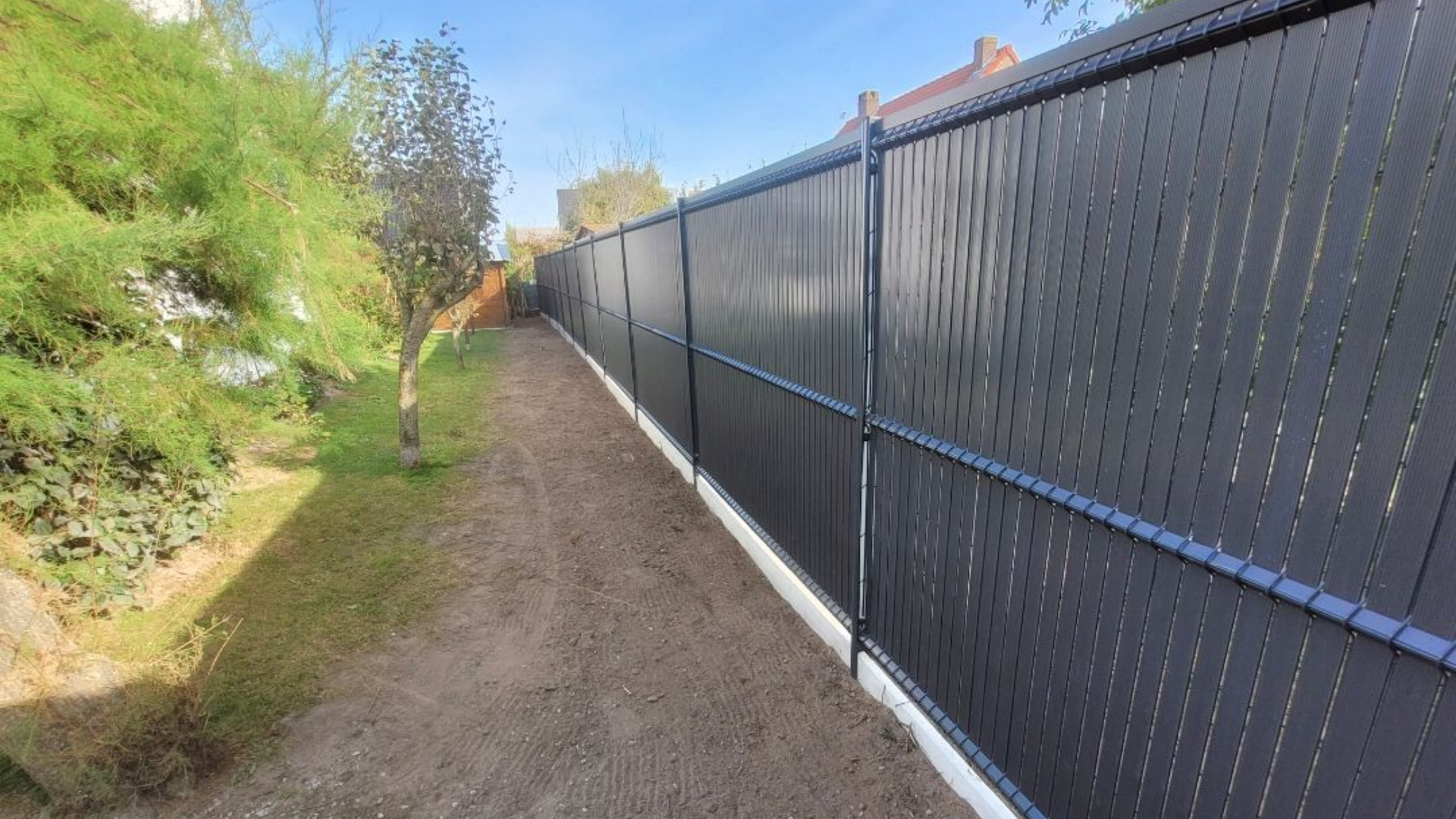Une clôture gris foncé borde un chemin de gravier, des buissons verts à gauche, un ciel bleu au-dessus.
