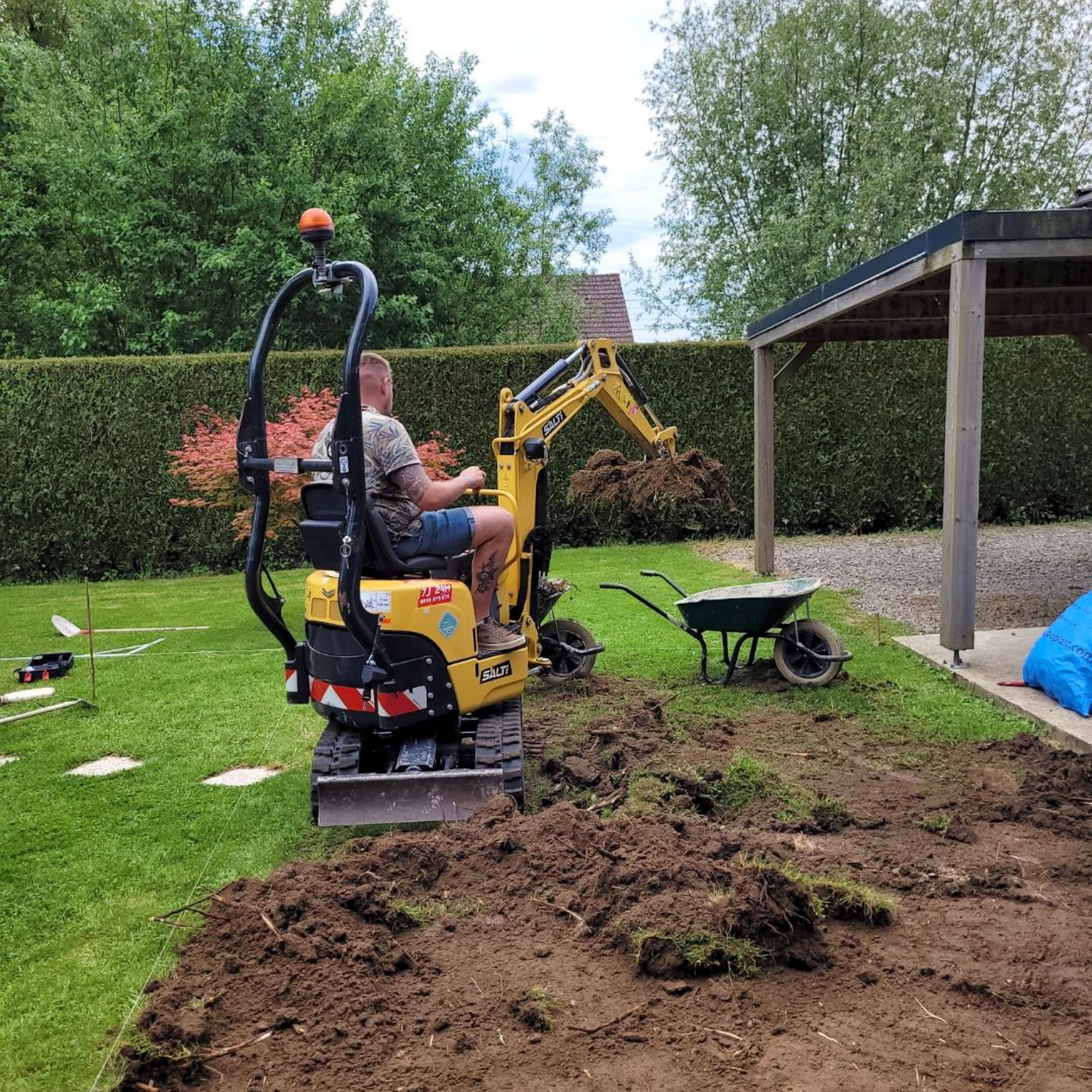 Un homme manœuvre une petite pelleteuse dans une cour, en train d'enlever de la terre. Une brouette se trouve à proximité.