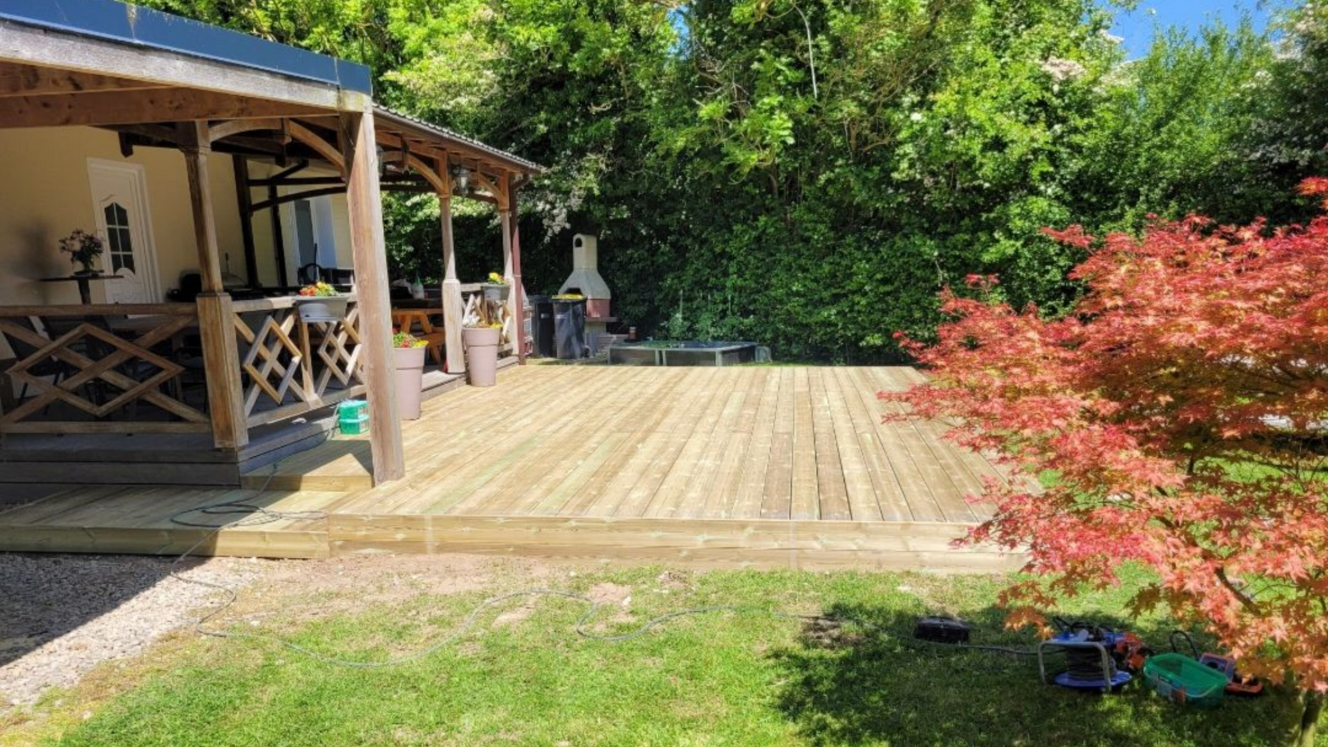 Terrasse en bois jouxtant un bâtiment, dans une cour gazonnée, avec un arbre rougeâtre et un feuillage vert.