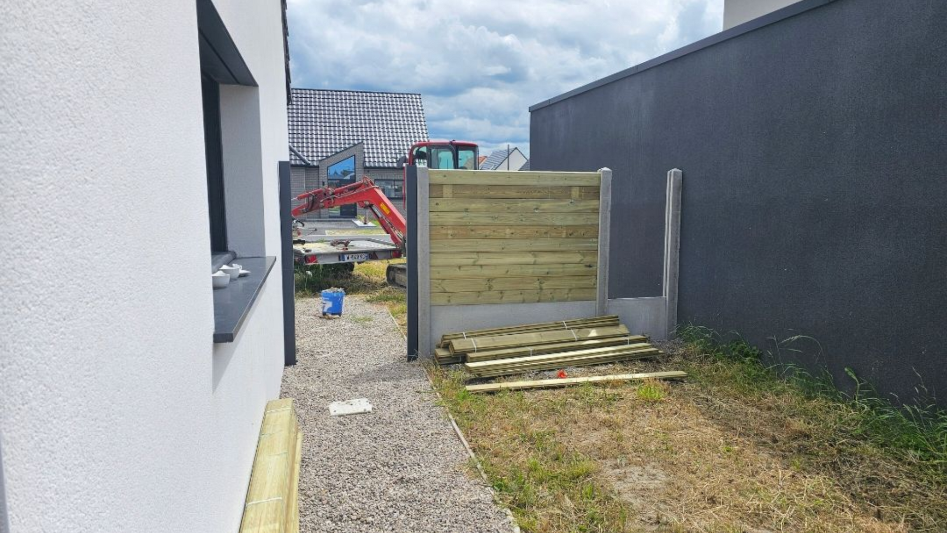 Clôture en bois avec chemin de gravier, côté d'un bâtiment blanc, excavatrice en arrière-plan.