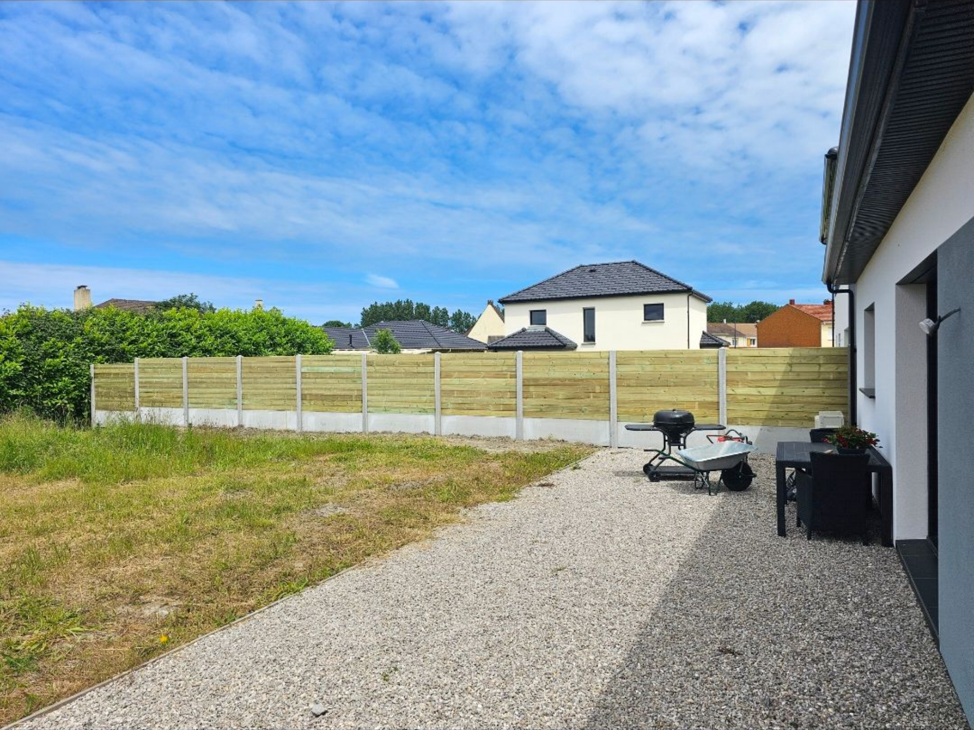 Vue sur le jardin : allée de gravier, pelouse, clôture en bois, façade blanche de la maison, ciel bleu.